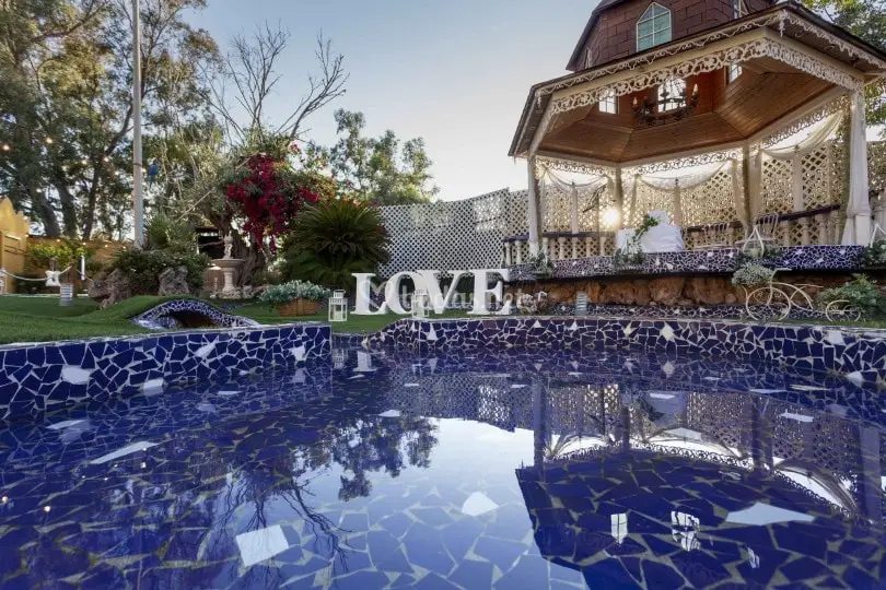 Un templete blanco de celosía y techo de madera sobre un estanque de mosaico azul, con las letras «LOVE» en el césped