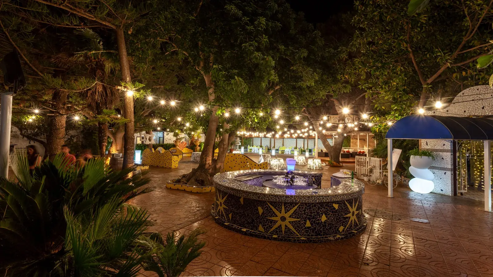 De noche, una fuente circular de mosaico con estrellas bajo árboles y guirnaldas de luces