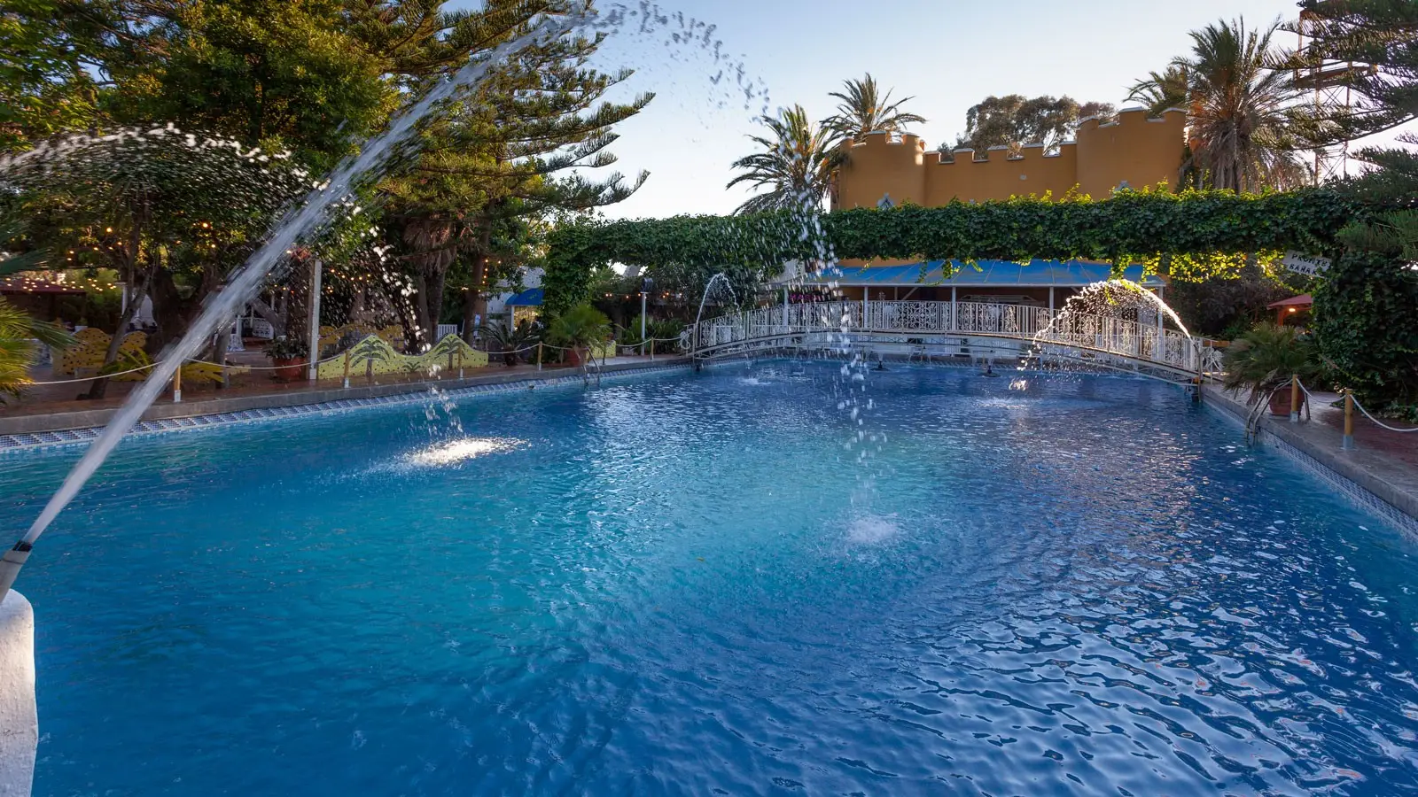 Una gran piscina con surtidores, un puente blanco al fondo y palmeras, con la terraza almenada amarilla de la masía
