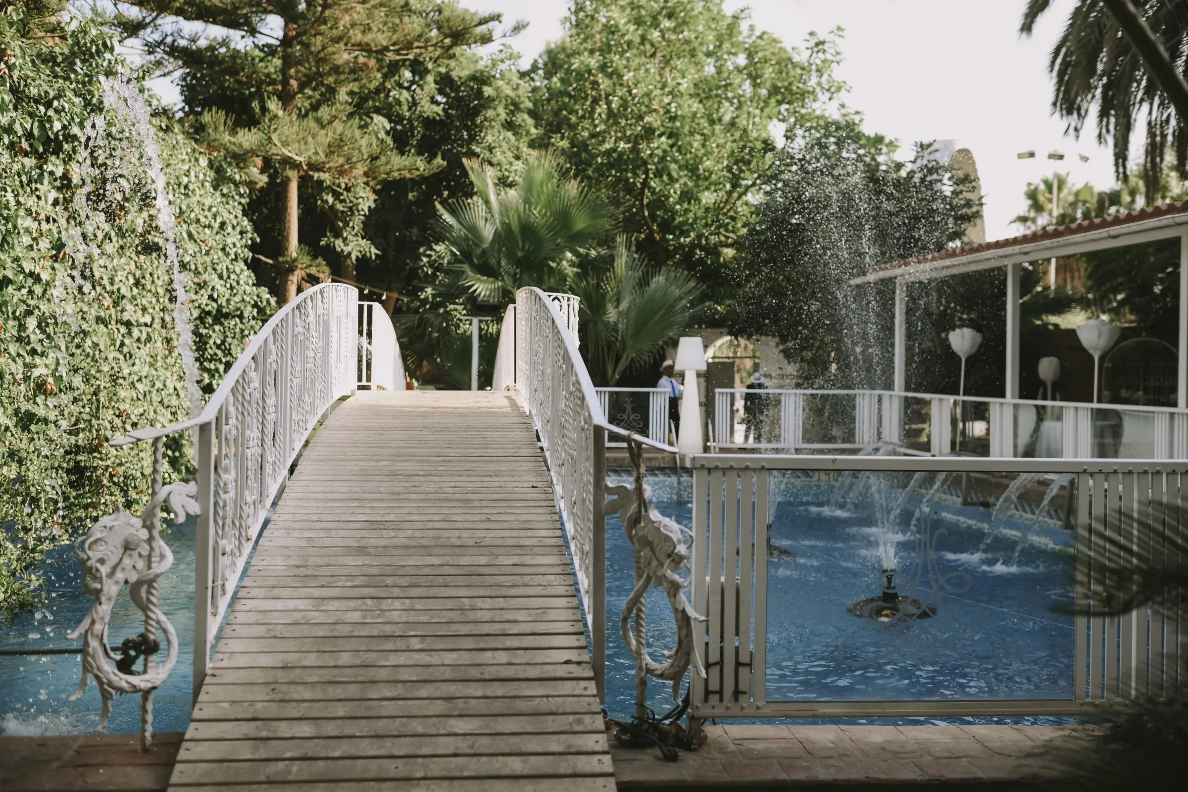 El puente de madera con barandilla blanca sobre el agua, con una fuente, pinos y palmeras